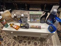 Cleaning supplies lot on white table including borax box, WD-40 can, spray bottles, caulking tube, wood oil, cleaners.