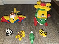 Photo shows the two toys with four animal figures on the floor: yellow Junny Jungle airplane and green Wow Toys Harry Copter helicopter with pilot figures. Toys are colorful and designed for toddlers.