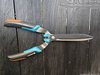 Side view of Gardena hedge shears showing ergonomic black handles with blue and orange rubber grips and long pointed steel blades.