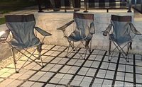 Three gray foldable outdoor chairs with black accents, each with built-in cup holders and side pockets, arranged on a tiled patio.