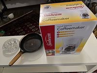 Photo showing the unopened box of Sunbeam 5-cup coffee maker, heavily used Vision pot, and heavy glass ashtray all placed on a white surface.
