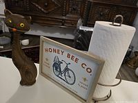 Photo showing the wooden cat sculpture, Honey Bee Co framed sign, and paper towel holder on a table with a paper towel roll on holder.