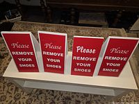 Four red and white foldable 'Please Remove Your Shoes' signs arranged on a white table surface showing front side text.