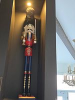 Full view of 48-inch life-size wooden nutcracker statue with red and blue painted details, standing in a niche against a dark wall. It wears a tall black fuzzy hat, holds a sword in one hand and a goblet in another, with detailed face and uniform.