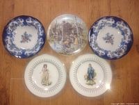 Five antique plates arranged on floor showing various designs including floral blue and white plates, village scene plate, and plates with historical figures.