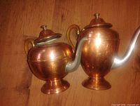 Two copper pots with wicker handles placed on a wooden floor, showing rounded bodies, metal spouts and lids.