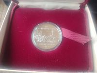 Photo of the silver dollar coin displayed inside a red velvet presentation box, showing the obverse and reverse sides through the capsule.