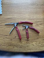 Photo of two E-ZRED Kiwi brand needle nose pliers with red handles on wood surface, showing the tool open and side profile.
