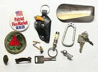 Overview of entire lot showing WWII military patch, Patriot Flea Market Dealer pin, various keys and keychains, tie clasp and pin, shoe horn and leather sheath with knife.