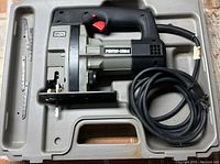 Top view of the jig/scroll saw inside the molded case with power cord coiled and several blades beside it.