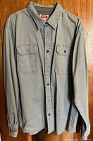 Full front view of Wrangler grey work shirt hanging on wooden background showing button-down front, collar, and two chest pockets.