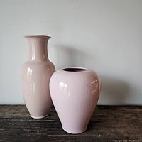 Both pink ceramic vases, one tall and one round side by side on wooden surface against white wall.