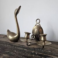 Both vintage brass pieces side by side on wooden surface, showing swan figurine and double candle sconce.