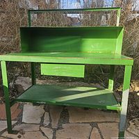 Green metal workbench outdoors on stone floor, showing overall structure, back guard, and shelves.