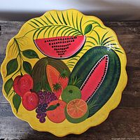 Top view of the round bowl showing colorful handpainted tropical fruits including watermelon, grapes, apples, lime, and orange on a yellow scalloped background.