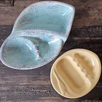 View of two ceramic ashtrays on wooden surface, showing blue speckled glaze ashtray with cigarette rests and yellow ashtray with crane design.