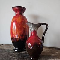 Two mid-century ceramic pieces placed side by side on a wooden surface against a white background: a tall, wide red lava glaze vase and a smaller red and brown pitcher with handle and spout.
