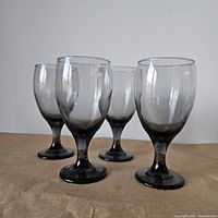 Four smoky grey tinted wine glasses placed on a flat surface, showing their shape and color gradient from clear top to dark stem and base.
