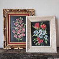 Two floral needlepoints displayed side by side with decorative frames on a wooden surface.