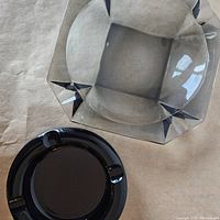 Photo showing both ashtrays on a paper surface, top-down view, highlighting the grey cut glass ashtray with geometric design and the smaller solid brown glass ashtray.