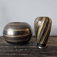 Photo of two vintage brass vases side by side on a wooden surface, one round and one tapered with banding designs