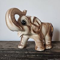 Side view of glossy ceramic elephant planter with raised trunk and visible detailed facial features, showing beige and brown coloring.