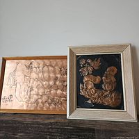 Two framed copper pieces leaning against a white wall on a dark wood surface: a copper map of Zambia with animals and an embossed copper squirrel artwork with a black background.