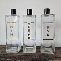 Front view of three vintage liquor bottles labeled Gin, Scotch, Rye with gold lettering and black plastic caps, arranged side by side on wooden surface