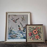 Both framed needlepoint artworks shown side by side against a white wall highlighting their relative sizes and colors.