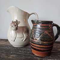 Two ceramic pitchers side by side on a rustic wooden surface; taller cream-colored with 3D cow design and smaller dark pottery pitcher with stripes.