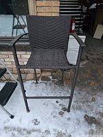 Front view of the outdoor bar stool showing woven seat and back, armrests, metal frame, and footrest on snowy ground outside a building.