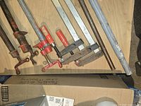 Top view of five metal pipe clamps with red handles laid on wooden surface, showing various clamp styles and some surface rust.