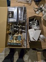 Box with multiple new door knobs, hinges, locksets, and door stops in original packaging taken from above.