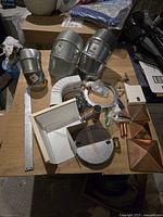 Top-down view of collection of new construction pipes, metal fittings, white plastic pipe elbow, eaves corner covers, and metal tape with other small plumbing materials on table.