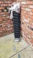 Black chainlink fence roll standing upright against brick wall, plastic wrapping on top, bound with ties.