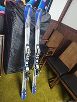 Pair of silver and blue Atomic M2tron skis standing upright with bindings attached, shown indoors against chairs and a black backdrop.