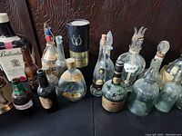 Photo showing a set of 15 antique and collectible bottles of various designs and sizes arranged on a black surface in front of a textured dark wall, displaying labels and bottle stoppers.