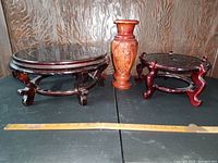 Two wooden raised plant stands and wooden Asian-style carved vase displayed on a black table against a wooden wall background with measuring stick for scale.