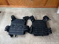 Two black tactical protective vests laying flat on a carpeted floor. Vests show adjustable straps and multiple MOLLE attachment points on the front.