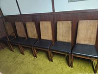 Six mid-century modern vintage dining chairs with cane backs and black upholstered seats shown lined up against wood-paneled wall on green carpet.