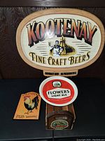 Photo of Kootenay Fine Craft Beers oval sign with Flowers Light Ale tray, Old Cock Pub rooster sign and wooden caddy.