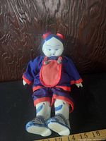 Full view of vintage Asian doll seated, showing ceramic head, hands, and feet with painted blue patterns and blue and red fabric clothing.