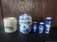 Photo of five blue and white vintage ceramic pieces: a cylindrical container with seascape, a lidded jar with scenic design, and three small blue vases with white floral or landscape motifs.