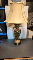 Full front view of the Bombay Company vintage table lamp with beige shade and ornate base on black surface.