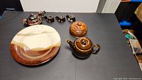 Overview photo showing the ceramic dragon teapot with three cups, the signed plate, another small brown teapot, and the carved wooden trinket box all arranged on a black surface.