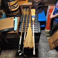 Full view of two vintage wood ski poles, curling broom, and hand-woven broom on a black surface surrounded by boxes and household items.