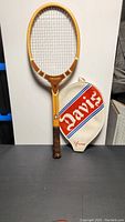 Full view of the vintage wooden Tad Davis tennis racket with attached strings and brown grip, next to the white case cover with red and blue 'Davis' lettering.