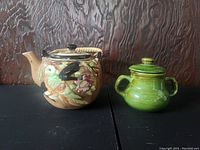 Photo of the vintage ceramic teapot with colorful bird and flower detail and woven handle next to green ceramic sugar bowl with lid and two handles on dark surface with wood textured background.