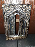 Black metal decorative door panel displayed hanging on wood surface, showing detailed floral and scroll relief work.