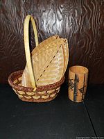 Woven basket holding 4 leaf-shaped woven trays next to a carved wooden vase, shown against a dark carved wood background.